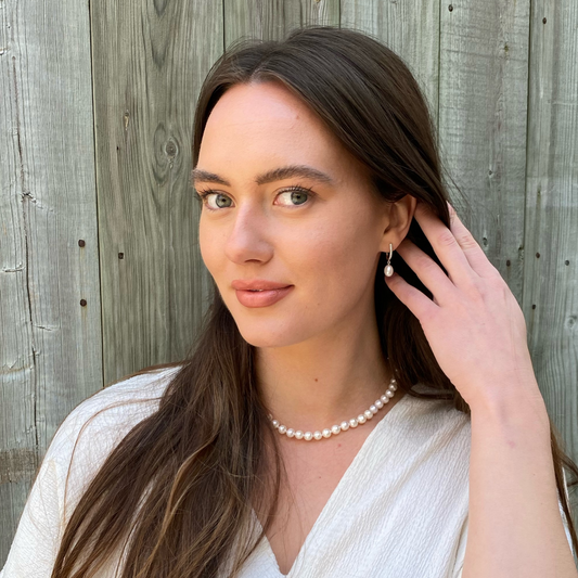 Woman with a classic pearl necklace adjusting her earring against a wooden fence background